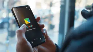En person håller en smartphone som visar Google Nyheter-appens logotyp och namn på skärmen, vilket föreslår att man kan ladda ner den från Google Play Butik, med en suddig utomhusscen synlig genom ett fönster i bakgrunden.