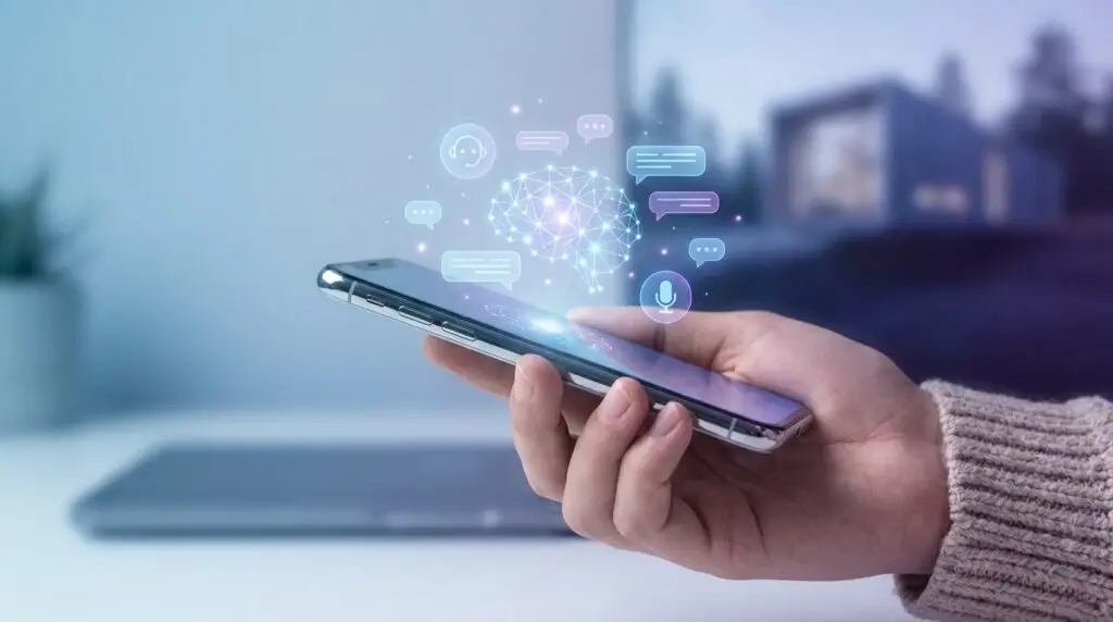 En person håller i en smartphone med holografiska ikoner av en hjärna, pratbubblor och en mikrofon ovanför, som representerar artificiell intelligens och digital kommunikation. En suddig laptop och ett hus syns i bakgrunden.