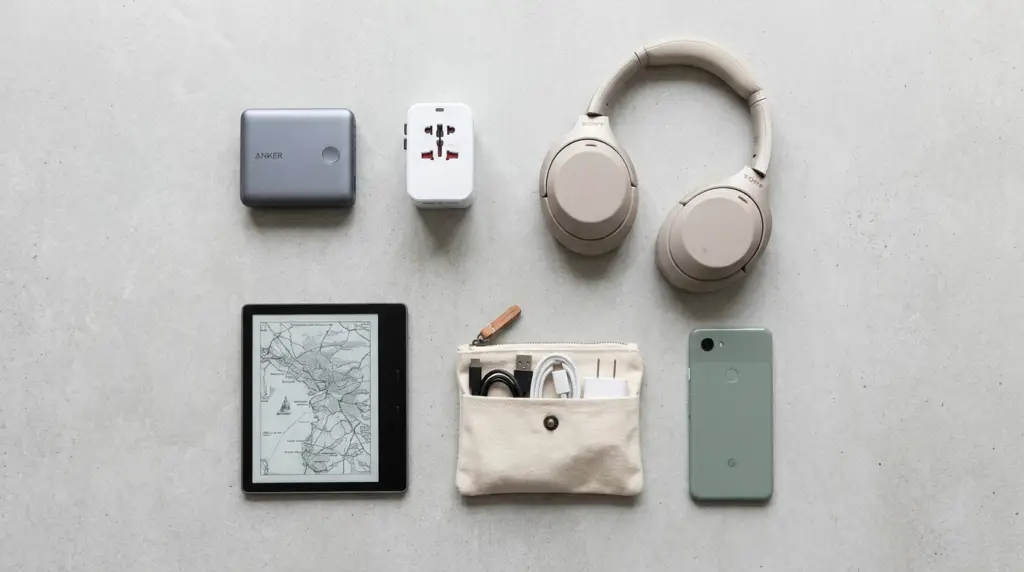 En plan bild av viktiga reseprylar på en grå yta, inklusive hörlurar, en powerbank, en universaladapter, en läsplatta med karta, en liten påse med kablar och en smartphone.