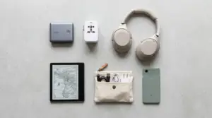 En plan bild av viktiga reseprylar på en grå yta, inklusive hörlurar, en powerbank, en universaladapter, en läsplatta med karta, en liten påse med kablar och en smartphone.
