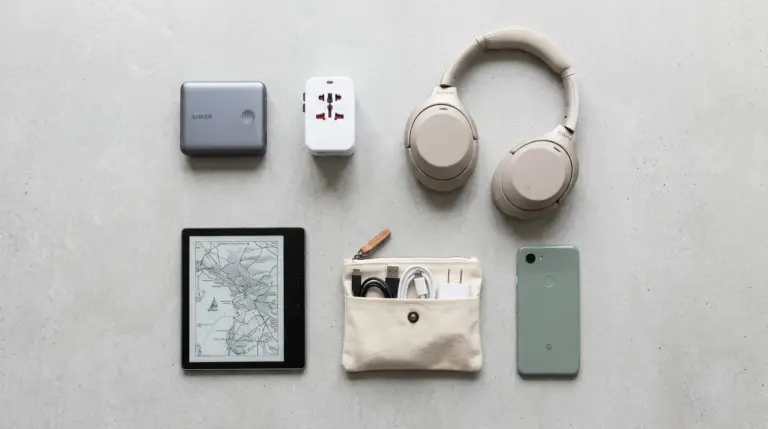 En plan bild av viktiga reseprylar på en grå yta, inklusive hörlurar, en powerbank, en universaladapter, en läsplatta med karta, en liten påse med kablar och en smartphone.